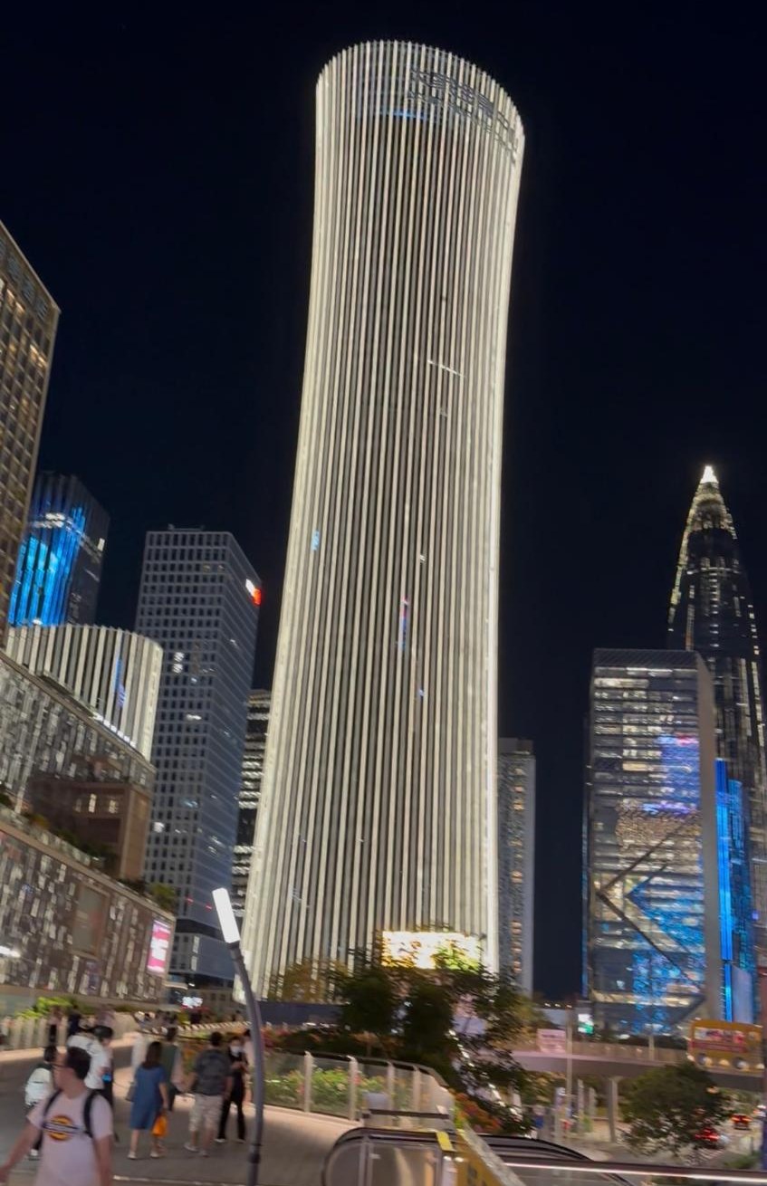 Shenzhen at night: a tall cylindrical tower lit in white stripes above a busy pedestrian walkway.