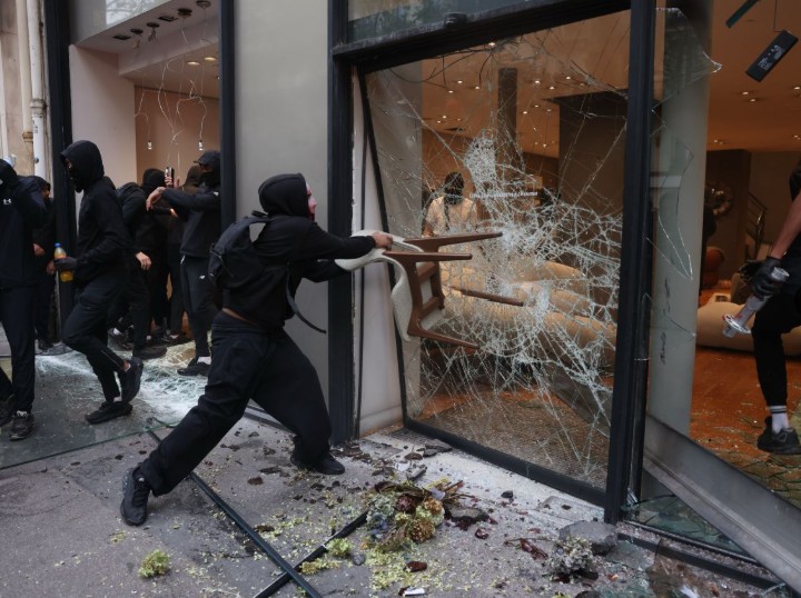 A masked rioter swings a chair into the shattered glass front of a luxury shop while others push inside.