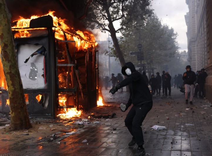 A bus shelter in flames on a Paris pavement, a masked youth about to throw a stone, a crowd behind him.