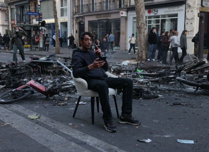 A man sits on an armchair in the middle of a Paris street among burnt-out bicycles, holding a phone.